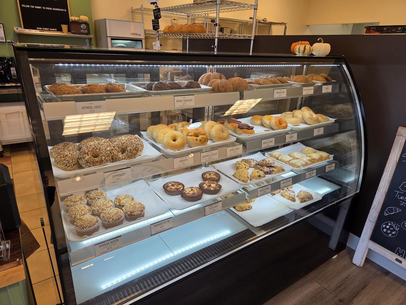 The pastry case: three shelves of cookies, bagels, scones and muffins with handwritten labels, and sourdough loaves on the rack behind.