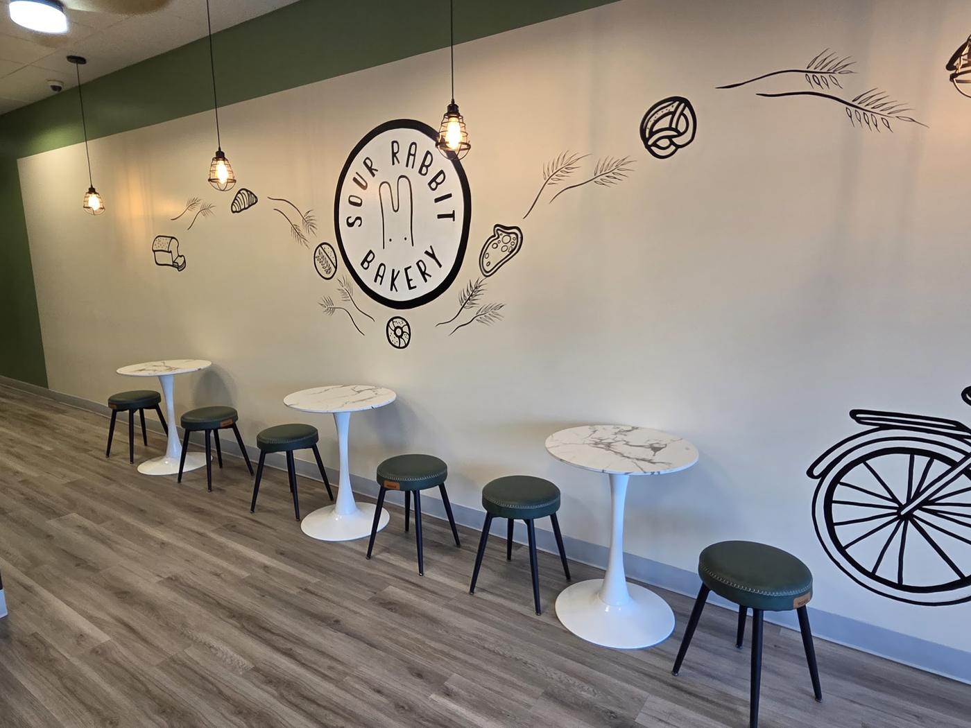 The bakery's interior feature wall: a hand-painted Sour Rabbit Bakery roundel surrounded by line drawings of bread, a croissant, a pretzel and wheat, with cafe tables and green stools beneath.